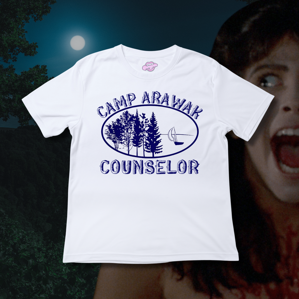 A classic white T-shirt with “Camp Arawak Counselor” and an illustrated navy blue camp logo featuring pine trees and a canoe. Background includes a spooky forest and a horror character screaming under the full moon.