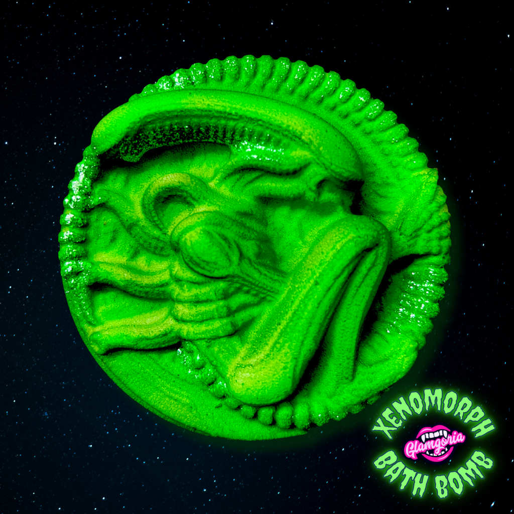 A vibrant green bath bomb shaped like the head of a xenomorph from the Alien franchise. The bath bomb features intricate details, including the creature's elongated skull and menacing facial structure, with ridges and curves that mimic the alien's iconic design. It is set against a dark, starry background, creating a cosmic and eerie atmosphere. The product name "Xenomorph Bath Bomb" and the Glamgoria logo are prominently displayed in the lower right corner.