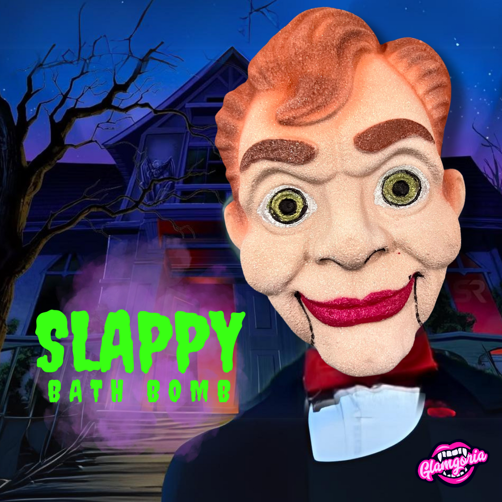 The image features a bath bomb designed to resemble Slappy, a character from the Goosebumps series. The bath bomb is in the shape of Slappy's face, showcasing his distinct features, such as his orange-brown wavy hair, large round eyes with dark circles, and a wide smile with red lips. The background shows a spooky house with bare trees against a dark, ominous sky, giving off a haunted vibe. The words "Slappy Bath Bomb" are written in a bright, dripping green font, with the "Glamgoria" logo in the bottom rig