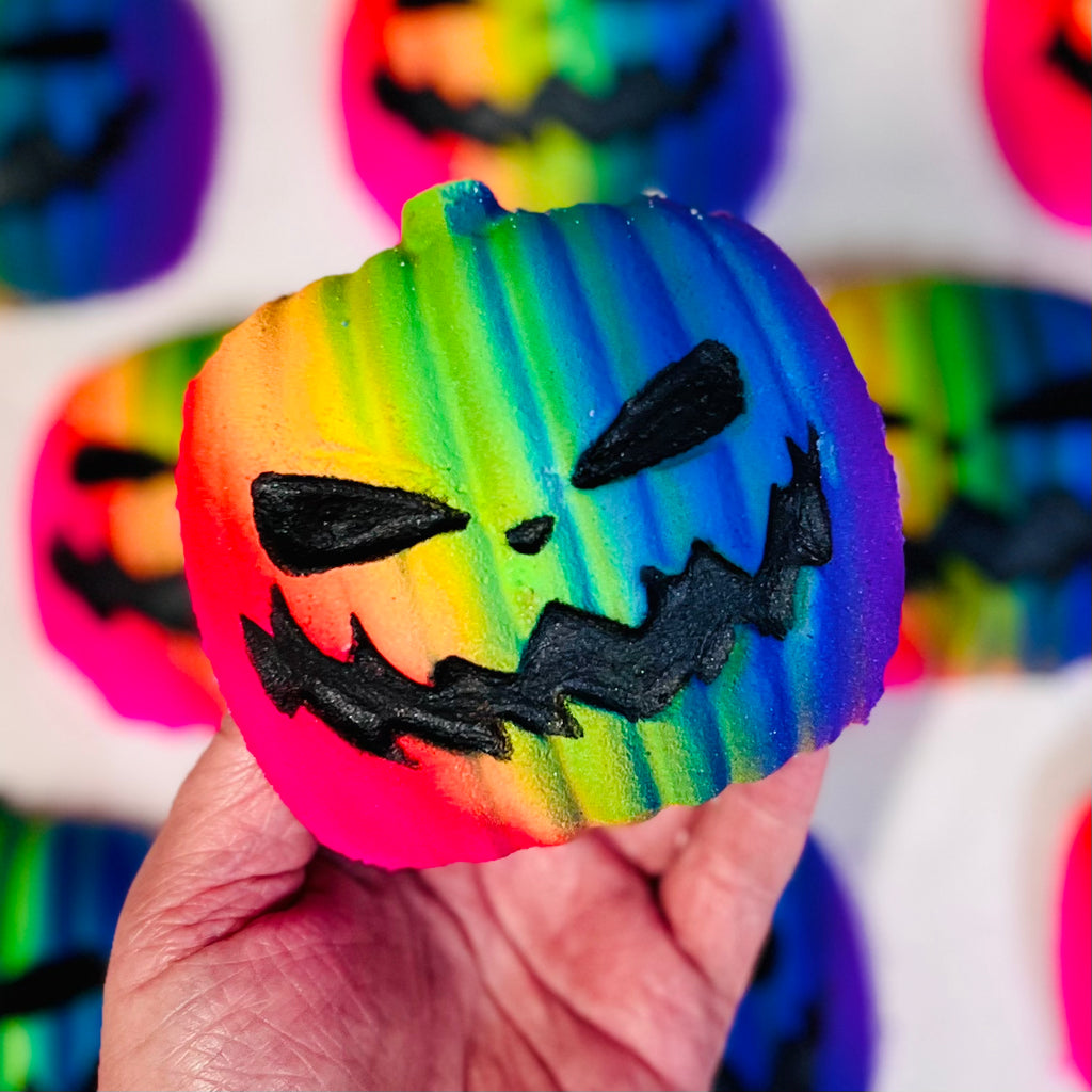 Hand holding a brightly colored neon rainbow pumpkin-shaped bath bomb with a black jack-o’-lantern face design, surrounded by other rainbow pumpkins in the background.