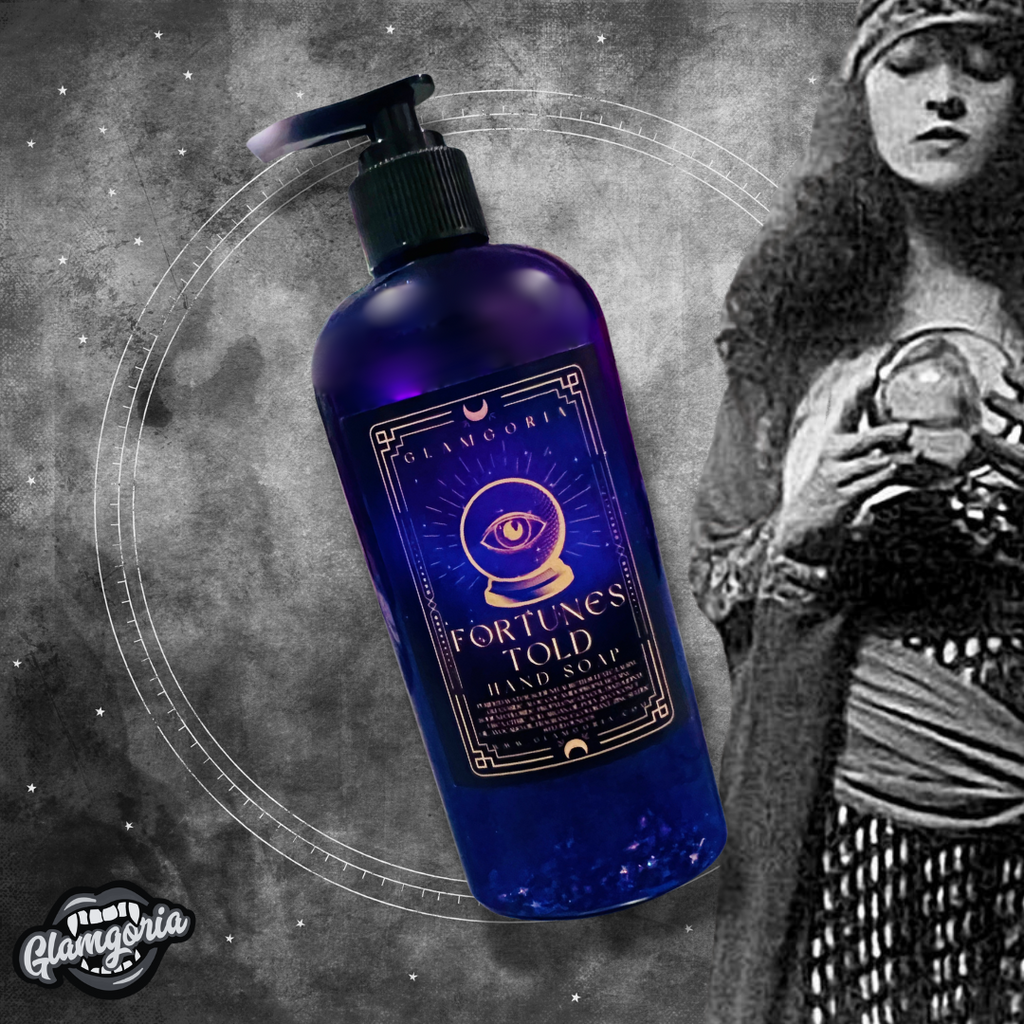 A mystic-themed image featuring the Fortunes Told shimmering hand soap in a deep purple bottle with an ornate label that displays a crystal ball and an eye. The soap has a subtle shimmer within the bottle. The background is a smoky grey with starry accents, and on the right side, there is a vintage black-and-white image of a fortune teller holding a crystal ball. The Glamgoria logo is placed in the bottom left corner."