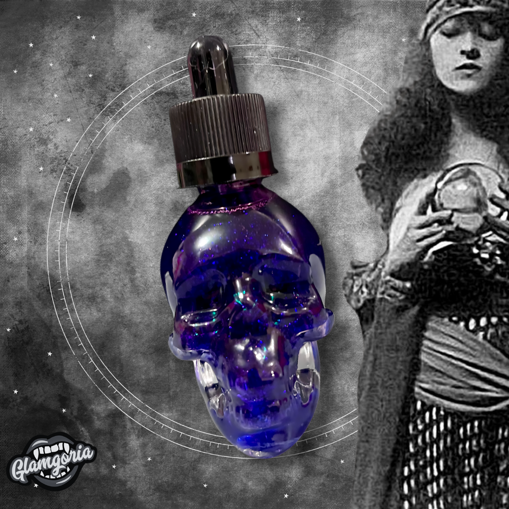 A stylized image featuring the Fortunes Told shimmering bath and body oil in a skull-shaped bottle with deep purple liquid and sparkling shimmer inside. The background is a mystical, smoky grey with starry accents, and on the right side, there is a vintage black-and-white image of a fortune teller holding a crystal ball. The Glamgoria logo is placed in the bottom left corner."