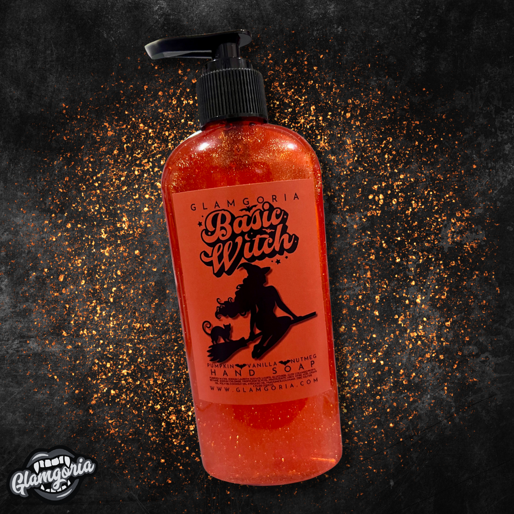 A bottle of Glamgoria’s *Basic Witch* hand soap with a rich, orange hue and shimmering sparkles inside. The label features a silhouette of a witch with flowing hair, sitting on a broomstick with a cat. The label text reads "Pumpkin, Vanilla, Nutmeg Hand Soap." The background is a dark, textured surface with orange sparkles scattered around the bottle.
