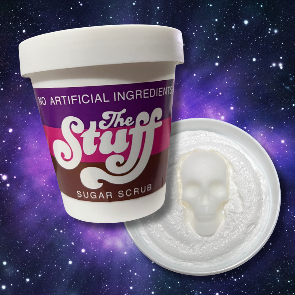 A horror-themed foaming sugar scrub inspired by The Stuff movie, featuring a white tub with a retro purple, pink, and brown label reading “The Stuff Sugar Scrub.” The scrub itself is white and fluffy with a skull-shaped soap embed in the center, set against a cosmic galaxy background.
