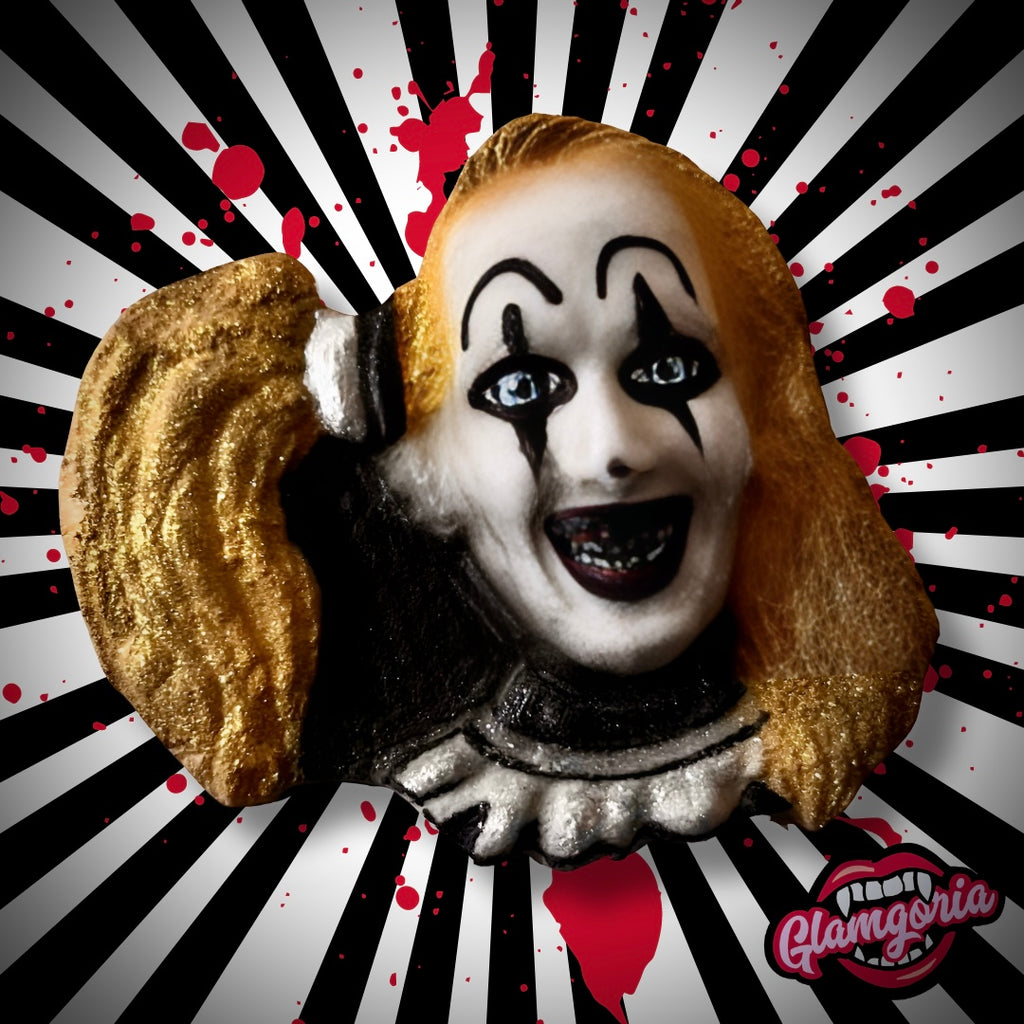 An eerie bath bomb shaped like the face of the Little Pale Girl from *Terrifier 2*, featuring detailed facial makeup with black tear-shaped designs under her eyes, a wide, unsettling smile, and golden hair. The background has black and white radial stripes with splashes of red, enhancing the creepy, horror-themed aesthetic. The Glamgoria logo, with a stylized set of fanged lips, is positioned in the lower right corner.