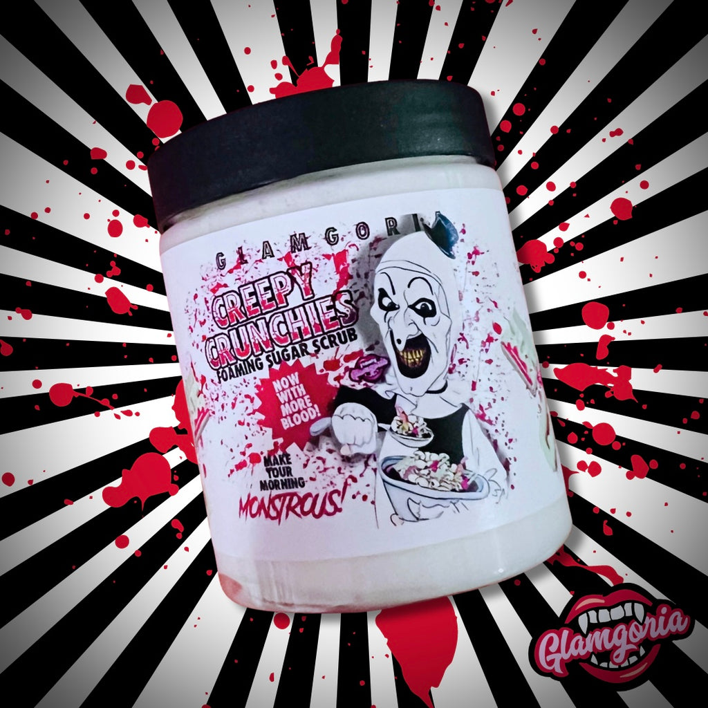 A jar of foaming sugar scrub labeled "Creepy Crunchies" by Glamgoria. The label features an illustration of a creepy clown character holding a bowl of cereal, with the tagline "Now with more blood!" The background of the image has a black and white radial pattern with red blood splatters. The jar is positioned slightly tilted, showcasing the label prominently.
