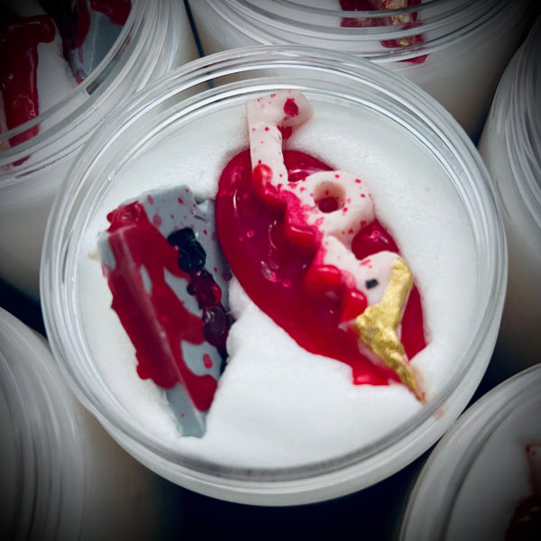 A close-up of the contents inside the jar of foaming sugar scrub. The scrub is white with embedded decorative pieces on top, including a bloody razor and corpse made of soap with red coloring to resemble blood. adding to the gory and horror-themed design of the product.
