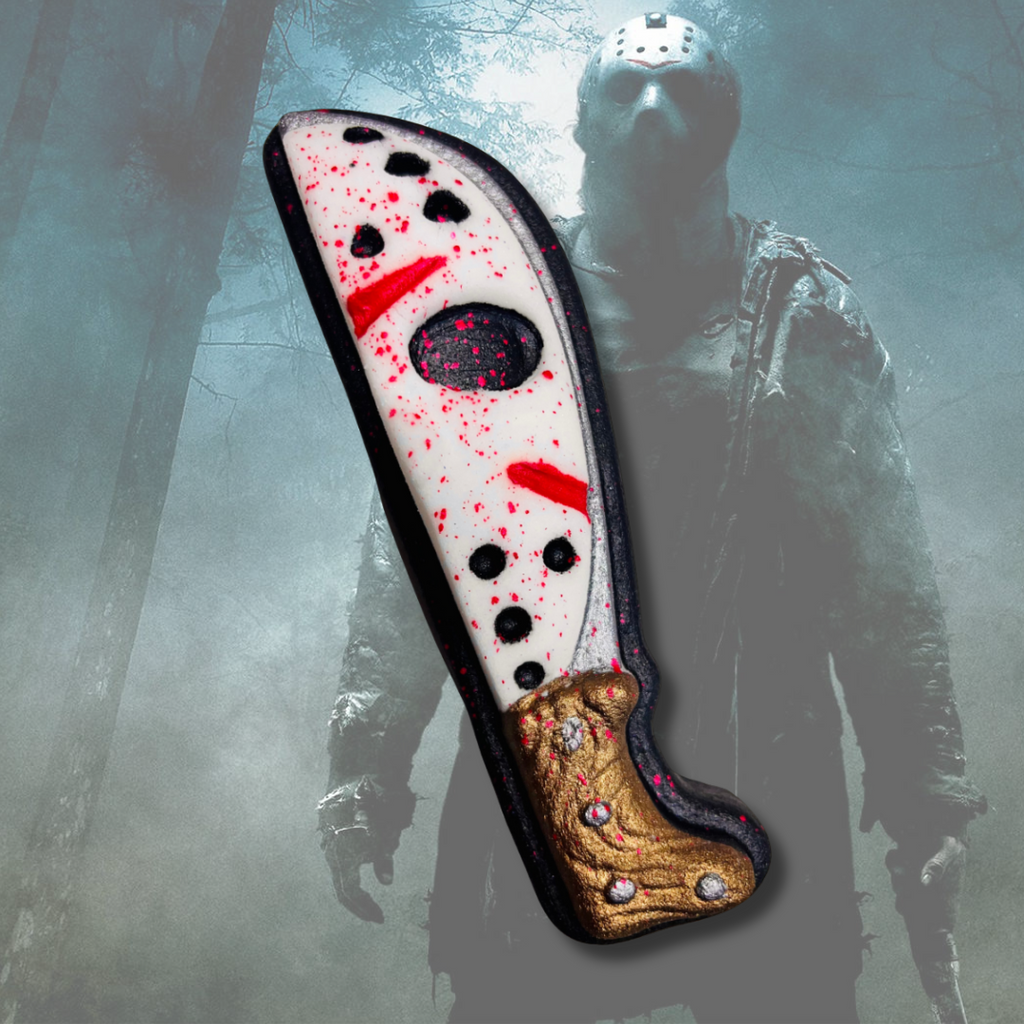 Machete-shaped bath bomb with white, black, and metallic gold details and red splatter accents, inspired by slasher horror and campground terror themes.
