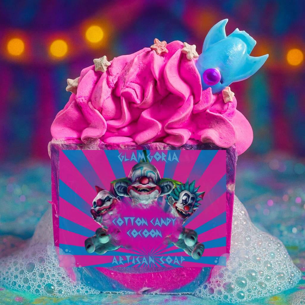 Bright neon-pink artisan soap topped with fluffy whipped soap frosting, pastel star embeds, and a blue cocoon-shaped topper. The label features three creepy clown illustrations on a pink and blue striped background with the text “Cotton Candy Cocoon.” Soap sits in foamy bubbles with a colorful carnival-style backdrop.
