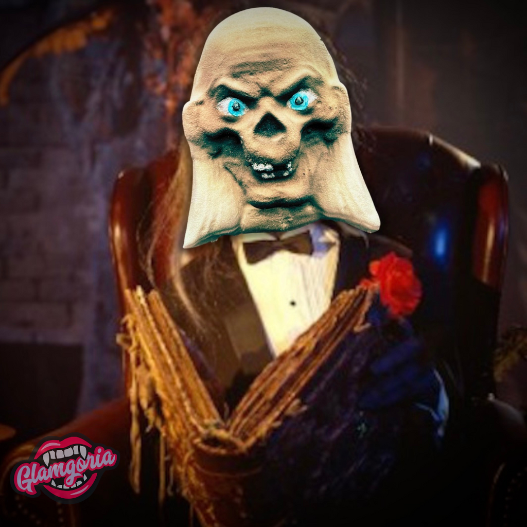 Cryptkeeper-inspired bath bomb styled as a skeletal ghoul with glowing blue eyes, dressed in a tuxedo and holding an old book, set against a dark, gothic background with Glamgoria logo.