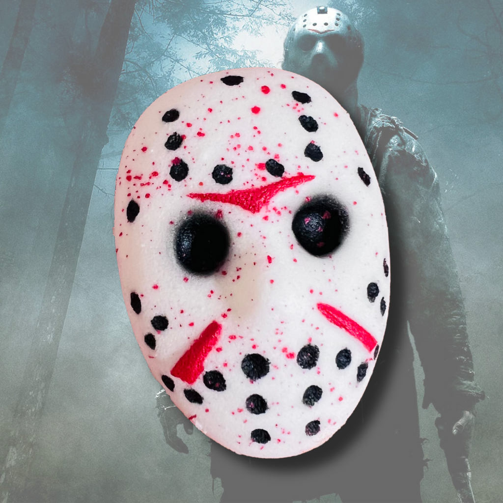 White hockey mask–shaped bath bomb with black detailing and red splatter accents, inspired by a slasher film aesthetic, set against a misty forest background.
