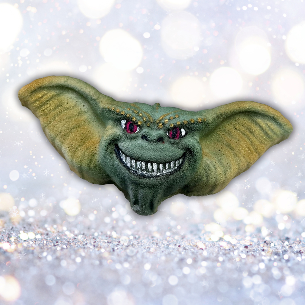Hand-painted green gremlin-themed bath bomb with wide ears and a mischievous grin on a sparkling winter background