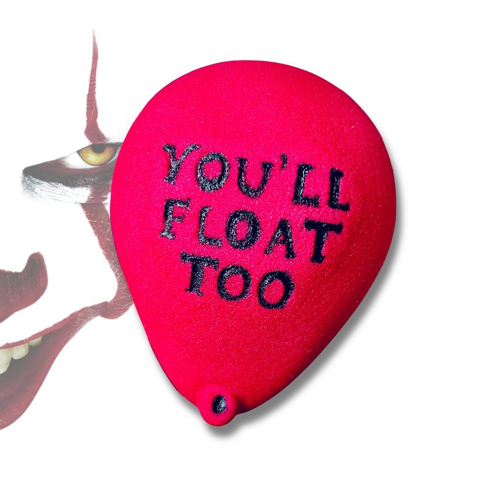 A red balloon-shaped bath bomb with the phrase “YOU’LL FLOAT TOO” written in black letters, referencing IT, on a white background.