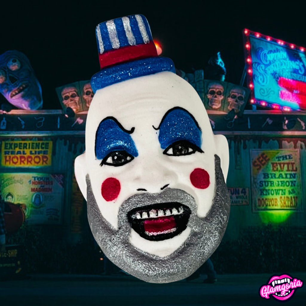 A bath bomb shaped like Captain Spaulding, the iconic clown character from the film *House of 1000 Corpses*, is shown. The bath bomb features Captain Spaulding's face with blue glittery eyeshadow, red cheeks, and a gray beard. His mouth is open in a wide grin, revealing glittery black teeth and red lips. He wears a small hat with red, white, and blue stripes. The background features a dark carnival scene with neon lights and creepy signs, adding to the horror-themed atmosphere. The lo