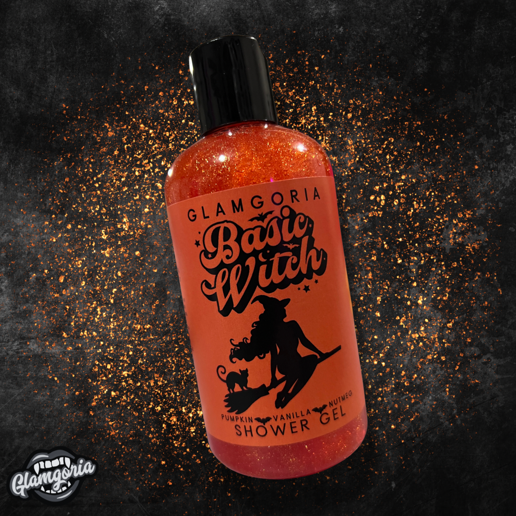 A bottle of Glamgoria’s *Basic Witch* shower gel with the same rich, orange color and shimmering sparkles. The label features a witch silhouette similar to the hand soap, with the text reading "Pumpkin, Vanilla, Nutmeg Shower Gel." The background is dark and textured, with orange sparkles scattered around the bottle.