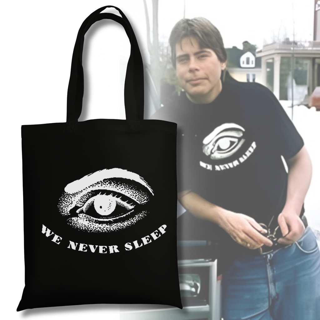 Black tote bag featuring a distressed illustrated eye with the phrase “We Never Sleep” printed beneath it in white.
