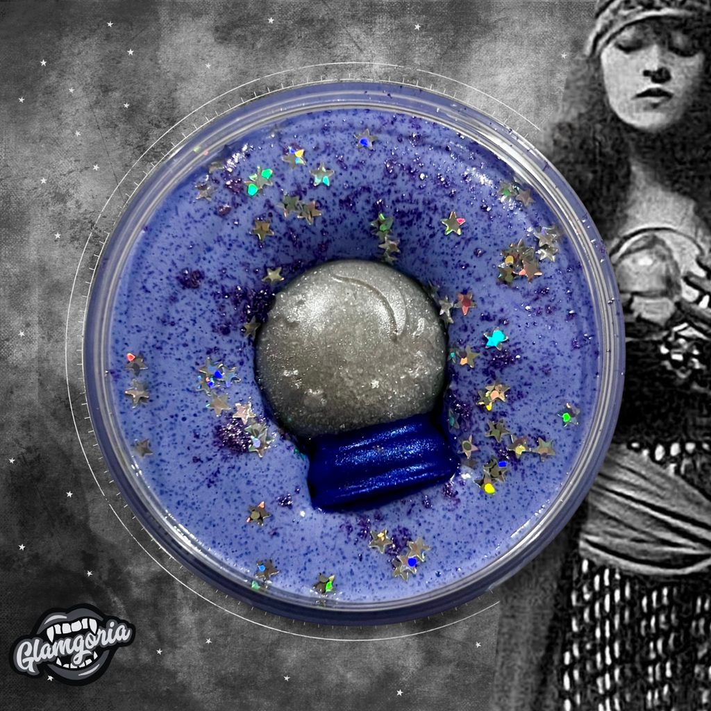 
"A top-down view of a foaming sugar scrub called 'Fortunes Told.' The scrub is a rich, deep purple color with a dusting of fine purple glitter and holographic star-shaped glitter scattered on top. In the center, there is a detailed, shimmering silver and blue snow globe soap topper embedded in the scrub, adding a magical and mystical touch."