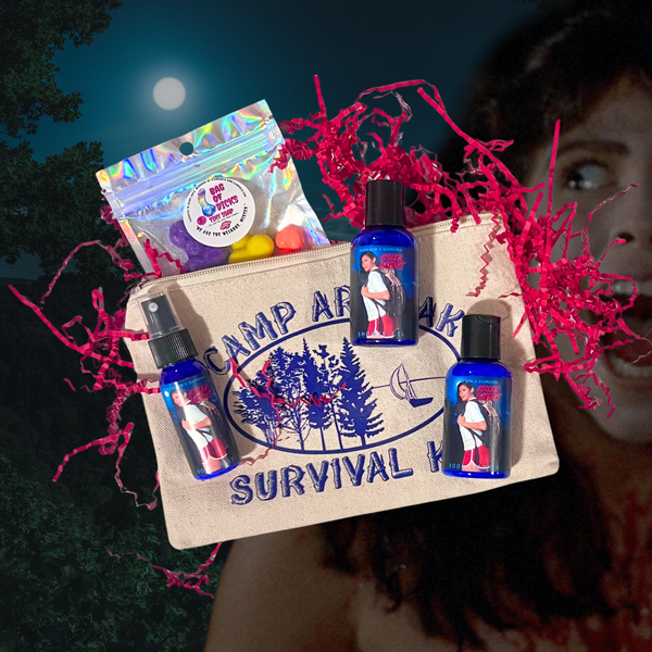 A horror-themed gift set featuring a canvas zip pouch labeled “Camp Arawak Survival Kit” surrounded by red crinkle paper. Includes blue bottles of Glamgoria shower gel, body lotion, body spray, and a holographic soap pouch labeled “Bag of Dicks.” Background includes a moonlit forest and Angela from Sleepaway Camp.