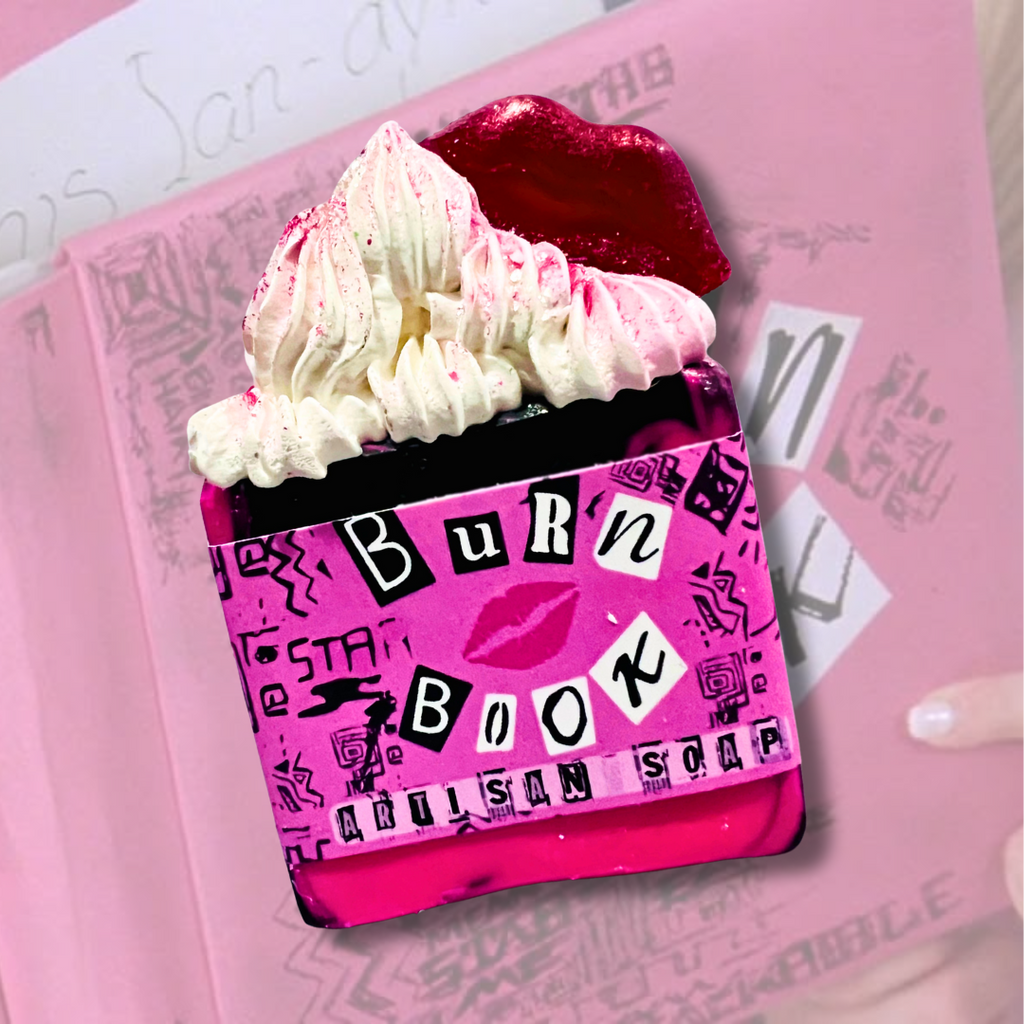 A vibrant hot pink artisan soap inspired by the Burn Book from Mean Girls, featuring whipped soap frosting, glittery red lips on top, and a cut-out style “Burn Book” label with doodles and lips. The background shows a real Burn Book page from the film. Pop culture-themed handmade soap for fans of iconic teen movies.