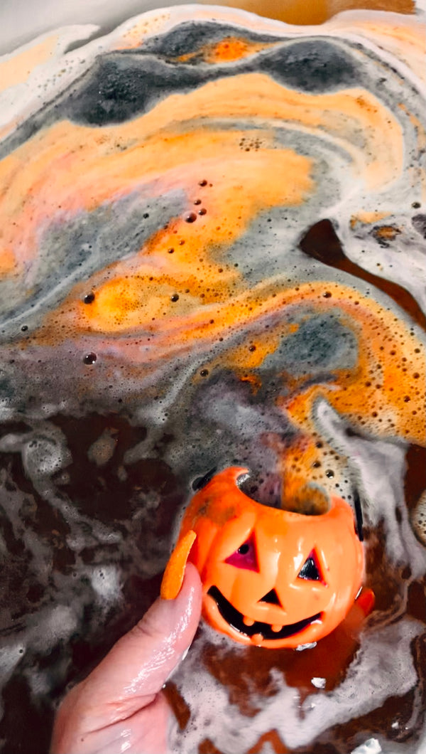 	A hand holding a Hack-o’-Lantern bath bomb floating in swirling orange and black bathwater.
