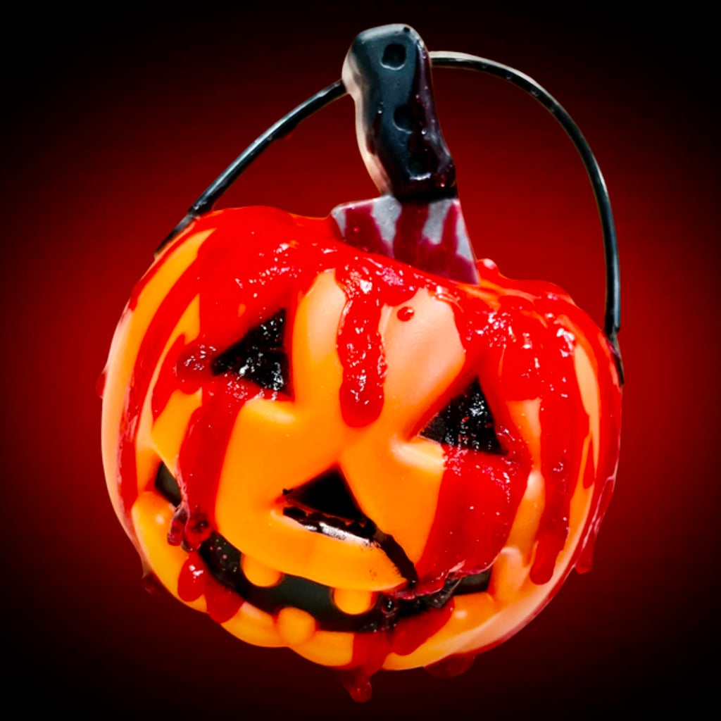 A small orange jack-o’-lantern bucket topped with a tiny butcher knife soap, dripping with realistic red “blood,” set against a dramatic red-and-black background.