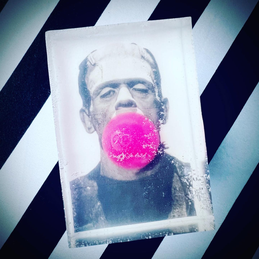 Clear rectangular soap with a black-and-white image of Frankenstein’s monster blowing a bright pink bubblegum bubble, set against a black and white diagonal striped background.

